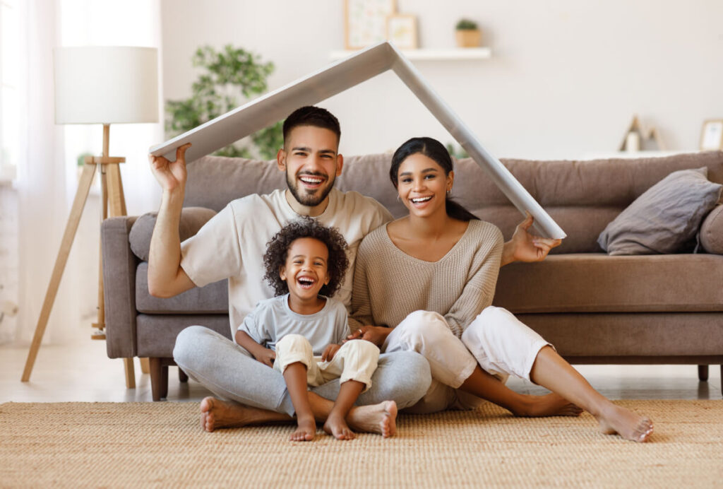 Cheerful parents with child smiling and keeping roof mockup over heads while sitting on floor in cozy living room